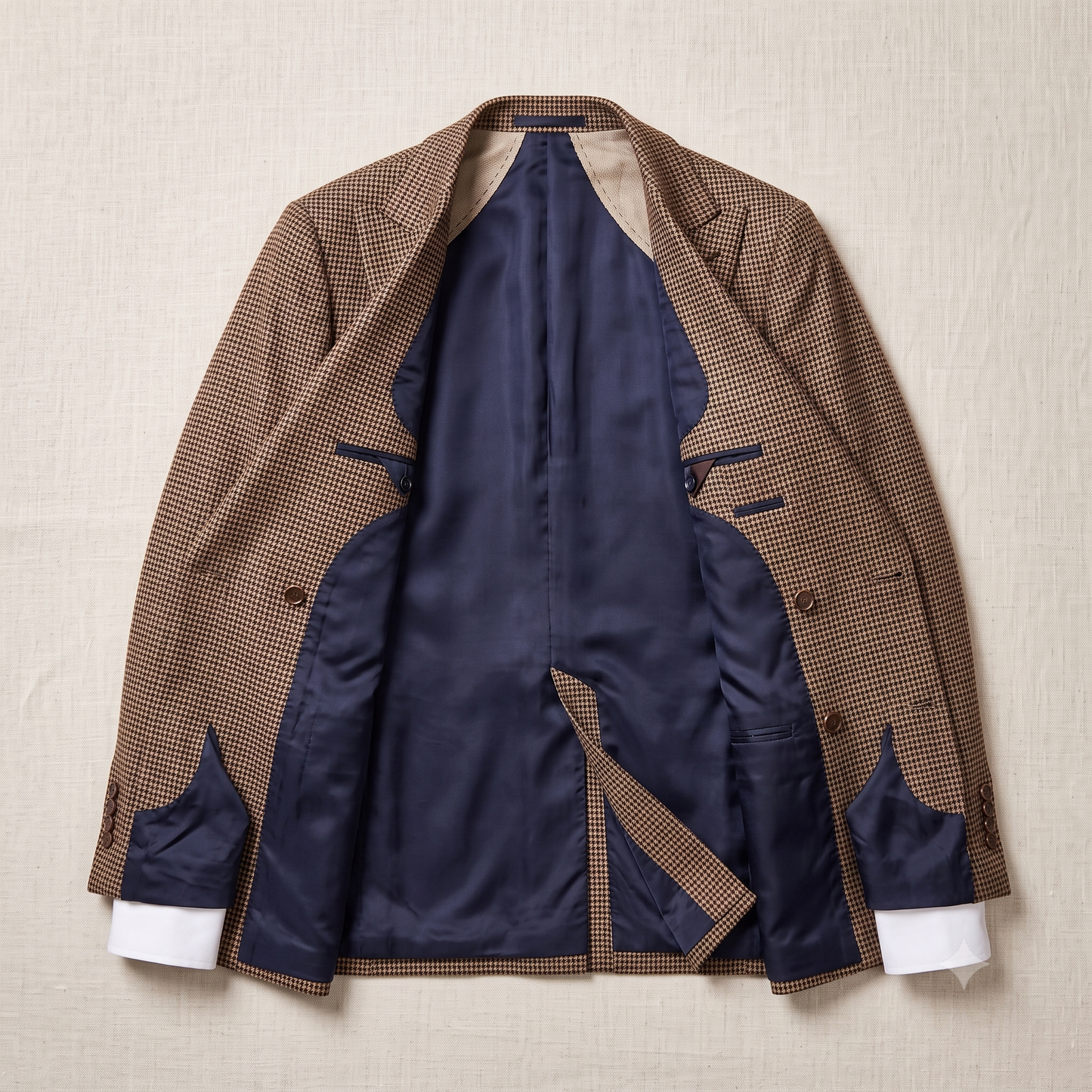 A brown and beige micro-check patterned blazer jacket is shown open, revealing a navy blue satin interior lining and white cuffs, laid flat on a textured beige fabric background.