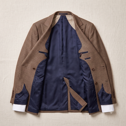 A brown and beige micro-check patterned blazer jacket is shown open, revealing a navy blue satin interior lining and white cuffs, laid flat on a textured beige fabric background.