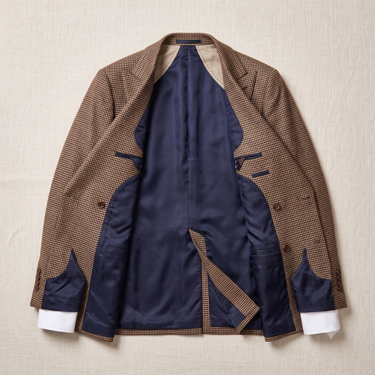 A brown and beige micro-check patterned blazer jacket is shown open, revealing a navy blue satin interior lining and white cuffs, laid flat on a textured beige fabric background.