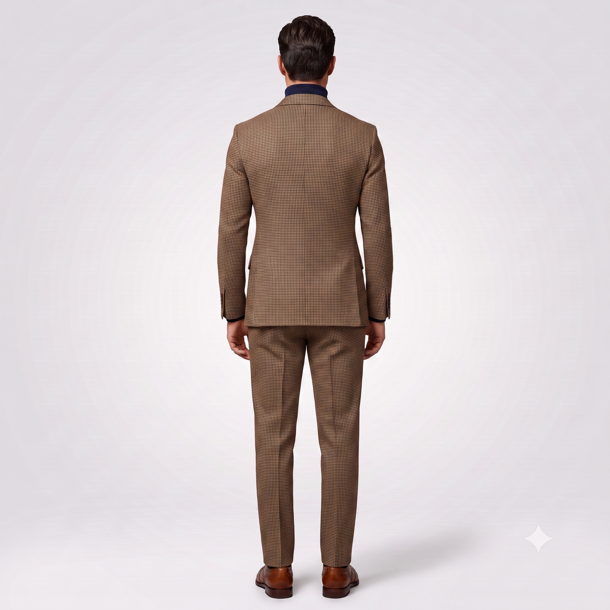 A man viewed from behind wearing a brown and beige micro-check patterned suit with a dark navy collar, brown dress shoes, and a white dress shirt, set against a light gray studio background.