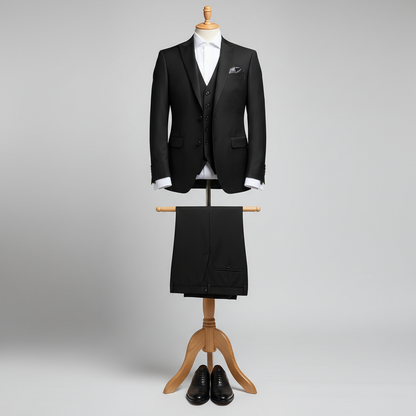 Three piece black men’s suit displayed upright on a wooden stand. Slim-fit jacket and matching waistcoat with clean tailoring, paired with coordinated trousers folded neatly over the display bar. Styled with a white dress shirt. Clean studio background with soft, even lighting. 250:250