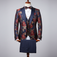 Three-piece men’s tuxedo displayed upright on a wooden stand. Slim-fit jacket crafted in deep navy fabric with bold red and gold floral jacquard pattern, finished with navy satin shawl lapels. Layered over a matching patterned waistcoat, paired with solid navy tailored trousers folded neatly over the display bar. Styled with a white dress shirt and matching patterned bow tie. Clean studio background with soft, even lighting highlighting the floral detailing and satin contrast.