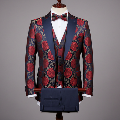 Three-piece men’s tuxedo displayed upright on a wooden stand. Slim-fit jacket crafted in deep navy fabric with bold red and gold floral jacquard pattern, finished with navy satin shawl lapels. Layered over a matching patterned waistcoat, paired with solid navy tailored trousers folded neatly over the display bar. Styled with a white dress shirt and matching patterned bow tie. Clean studio background with soft, even lighting highlighting the floral detailing and satin contrast.