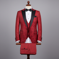 Three-piece red men’s tuxedo displayed upright on a wooden stand. Slim-fit jacket crafted in red jacquard fabric with subtle floral pattern, finished with black satin shawl lapels and a black chest pocket accent. Layered over a matching red waistcoat, with coordinated trousers folded neatly over the display bar. Styled with a white dress shirt and black bow tie. Clean studio background with soft, even lighting highlighting the pattern and satin contrast.