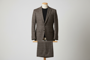 Two-piece brown men’s suit displayed upright on a wooden stand. Slim-fit jacket with structured shoulders and a defined check pattern formed by fine intersecting lines, paired with matching trousers folded neatly over the display bar. Styled with a black top for contrast. Clean studio background with soft, even lighting highlighting the fabric texture and line detailing.