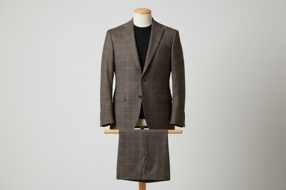 Two-piece brown men’s suit displayed upright on a wooden stand. Slim-fit jacket with structured shoulders and a defined check pattern formed by fine intersecting lines, paired with matching trousers folded neatly over the display bar. Styled with a black top for contrast. Clean studio background with soft, even lighting highlighting the fabric texture and line detailing.