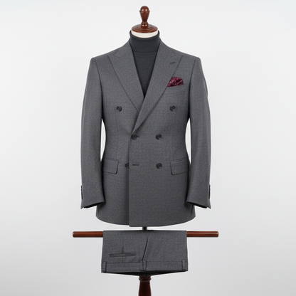 Two-piece grey men’s suit displayed upright on a wooden stand. Double-breasted slim-fit jacket crafted in a textured grey woven fabric with fine micro-pattern detailing, paired with matching trousers folded neatly over the display bar. Styled with a dark turtleneck and pocket square. Clean studio background with soft, even lighting highlighting the texture and structure.