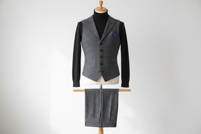 Two-piece men’s waistcoat set displayed upright on a wooden stand. Slim-fit check-pattern waistcoat with matching trousers folded neatly over the display bar. Styled with a black turtleneck for a modern contrast. Clean studio background with soft, even lighting.
